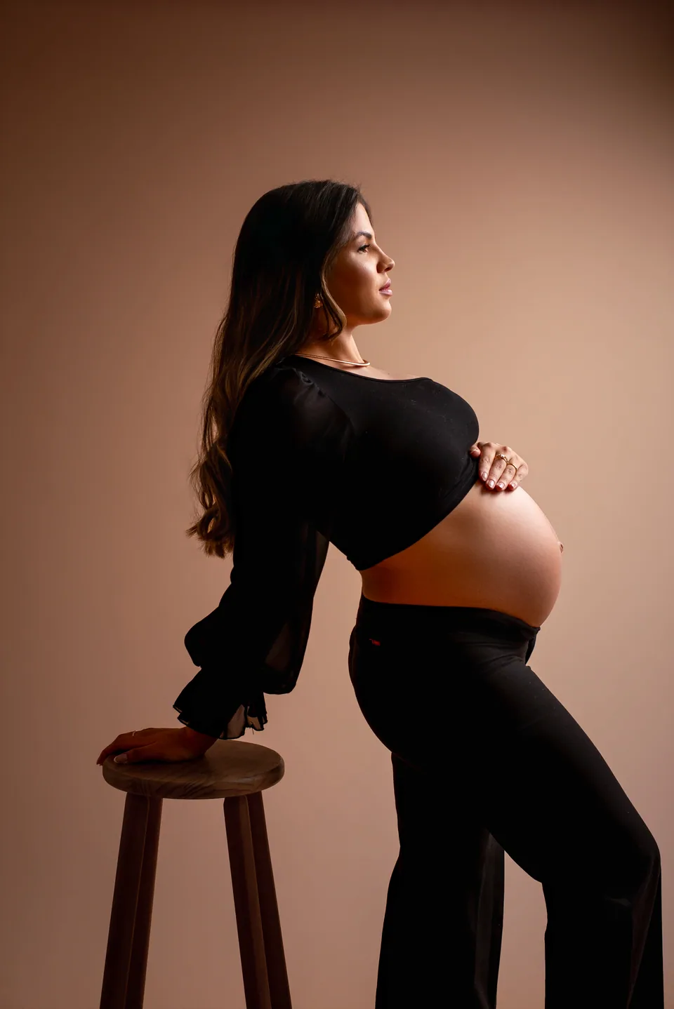Gestante de perfil em estúdio, blusa preta, mão sobre a barriga e apoio em banco de madeira sob luz quente