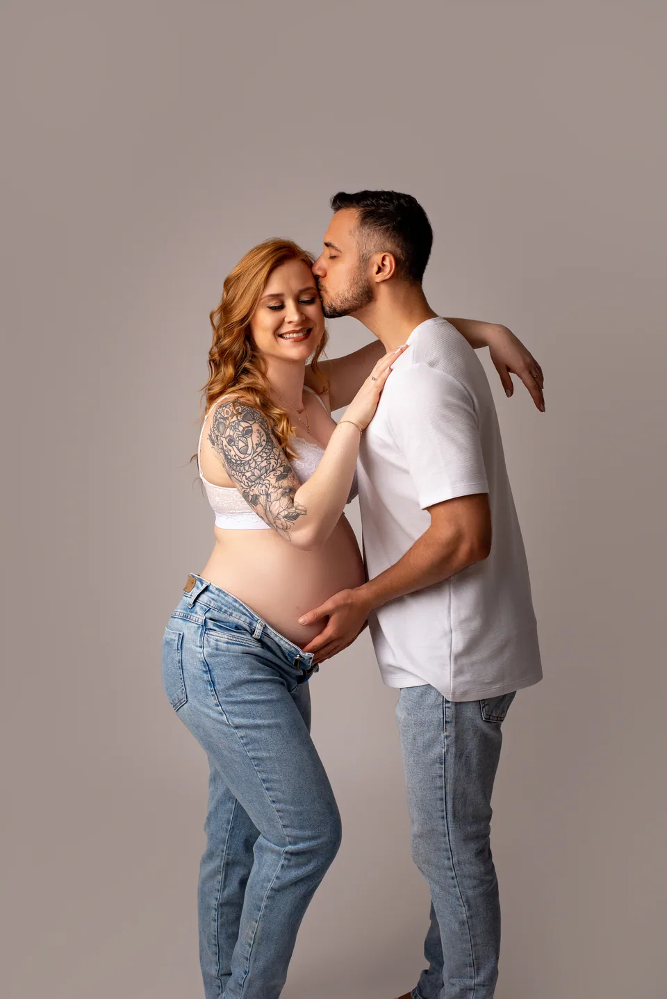 Casal em ensaio gestante: pai beija a testa da mãe, que sorri de olhos fechados, com a mão dele na barriga