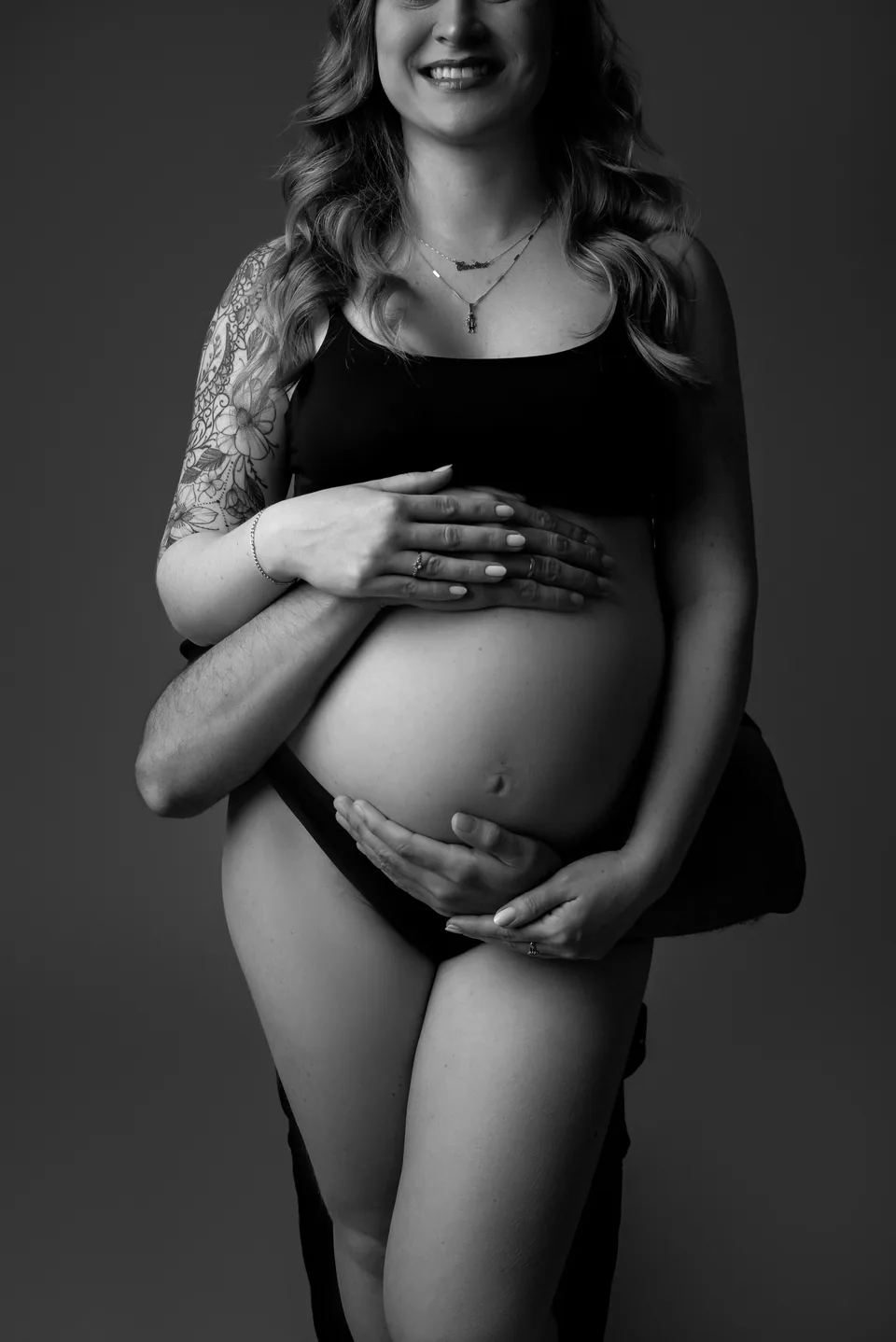 Ensaio gestante em preto e branco: mãe sorridente de top preto, quatro mãos envolvendo a barriga
