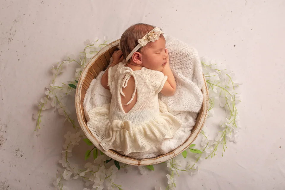 Recém-nascida dormindo em cesto de madeira, vestido de tricô creme, faixa de flores e flores brancas ao redor