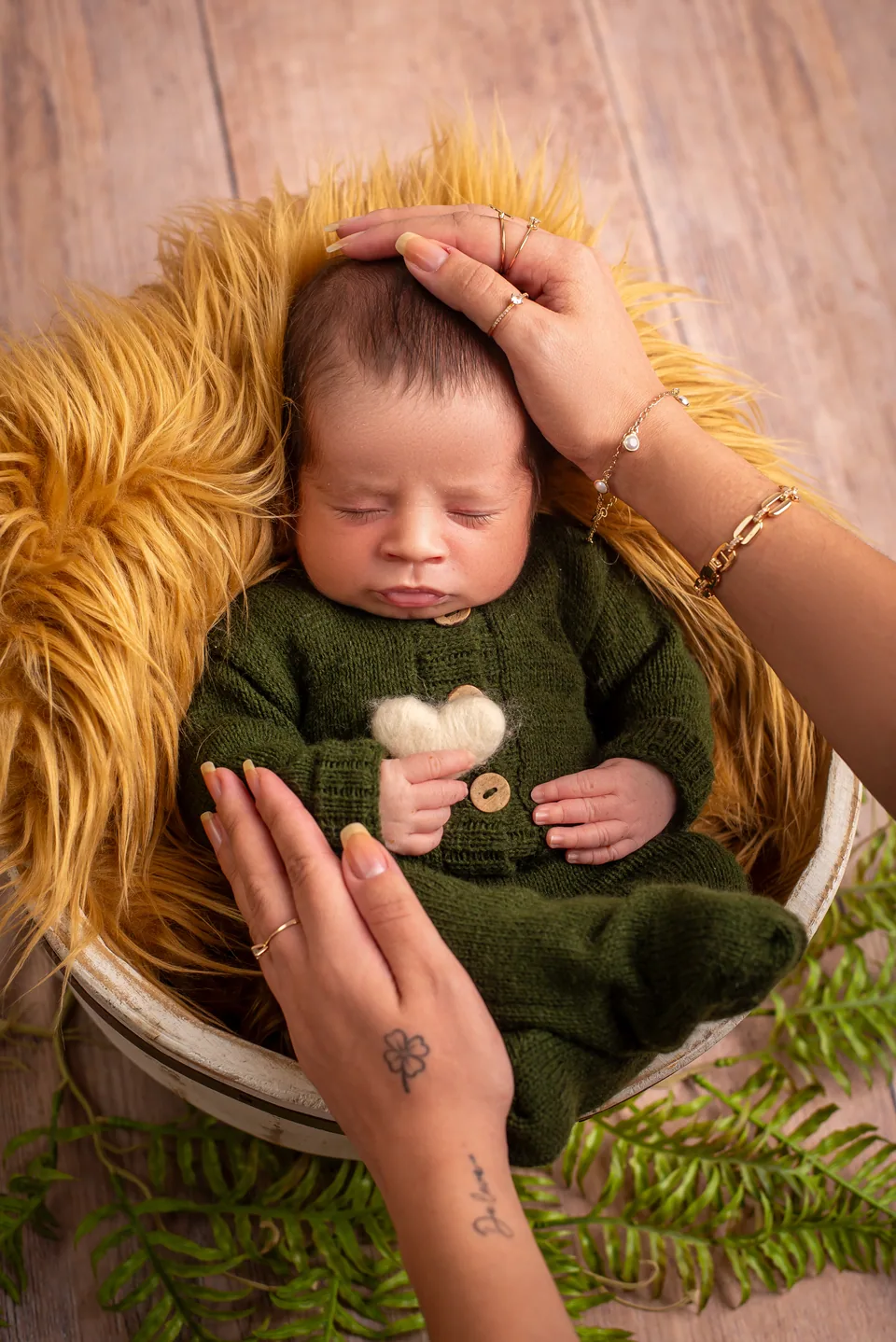 Recém-nascido de macacão de tricô verde com coração de feltro nas mãos, deitado em pelo amarelo, mãos da mãe apoiando